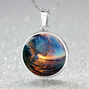 Op de afbeelding: Zilveren ketting met een ronde hanger met een kleurrijke foto van een golf die bij zonsondergang in de oceaan breekt.