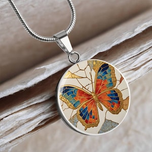 May include: A circular silver-toned pendant necklace featuring a stained-glass style butterfly design. The butterfly has vibrant orange, red, and blue segments with gold-colored outlines, set against a cream-colored background with cracked patterns.