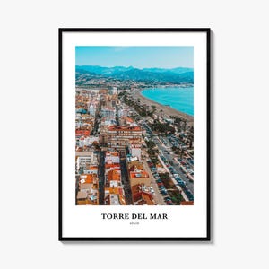 Könnte beinhalten: Ein gerahmter Kunstdruck mit einer Luftaufnahme der Küstenstadt Torre del Mar in Spanien. Das Bild zeigt einen Sandstrand, türkisfarbenes Wasser und eine dichte städtische Bebauung mit orangefarbenen Dächern unter einem klaren blauen Himmel.