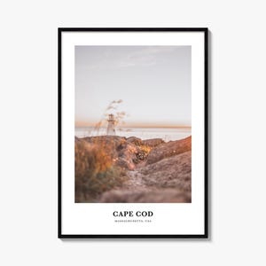 Könnte beinhalten: Ein gerahmter Kunstdruck mit einer Fotografie eines weißen Leuchtturms an einer felsigen Küste bei Sonnenuntergang. Im Vordergrund sind unscharfe goldene Gräser und strukturierte Felsen zu sehen. Der Text unten lautet CAPE COD MASSACHUSETTS, USA.