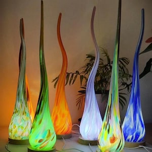 May include: Six colourful glass lamps with a unique, flowing design. Each lamp has a different colour and pattern, including green, blue, purple, orange, and white. The lamps are on wooden bases and have a warm, inviting glow.