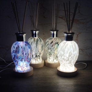 Peut inclure: Quatre flacons diffuseurs en verre avec des bases en bois et des roseaux. Les flacons sont éclairés de l'intérieur, créant une lueur chaleureuse. Les flacons sont décorés de motifs différents, notamment du blanc, du bleu et du vert.