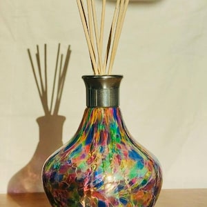 Puede incluir: Una botella de difusor de vidrio colorida con una base de madera y cañas. La botella tiene una tapa plateada y está llena de un arcoíris de colores.