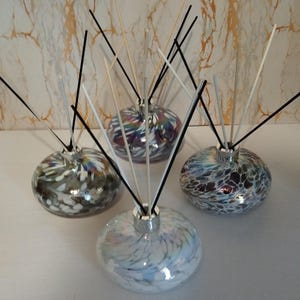 Może przedstawiać: Cztery dekoracyjne szklane dyfuzory do aromaterapii z patyczkami. Okrągłe szklane butelki mają kolorowe, wirujące wzory w odcieniach niebieskiego, fioletowego i białego. Patyczki są czarne i naturalne, tworząc nowoczesną estetykę.