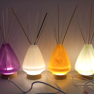 May include: Four decorative aroma diffusers with glass teardrop-shaped containers in purple, white, orange, and green. Each diffuser has wooden bases and a bundle of reeds. The diffusers are lit from within, creating a soft glow.