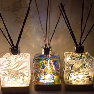 Könnte beinhalten: Drei Glasdiffuserflaschen mit Holzsockeln und schwarzen Diffusorstäbchen. Die Flaschen sind quadratisch und haben unterschiedliche Muster: Weiß mit Wirbeln, mehrfarbig und Weiß mit einem Blumenmuster. Die Flaschen sind von innen beleuchtet und erzeugen ein warmes Leuchten.