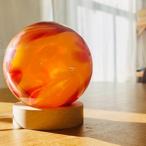 Peut inclure: Un orbe de verre rond, orange et rouge avec des motifs tourbillonnants repose sur une base en bois. L'orbe est éclairé de l'intérieur, créant une lueur chaude.
