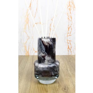 May include: A marbled black and grey reed diffuser with white reeds. The cylindrical glass vessel sits on a clear base. The background features a gold and white marble pattern. The diffuser is designed for home fragrance.
