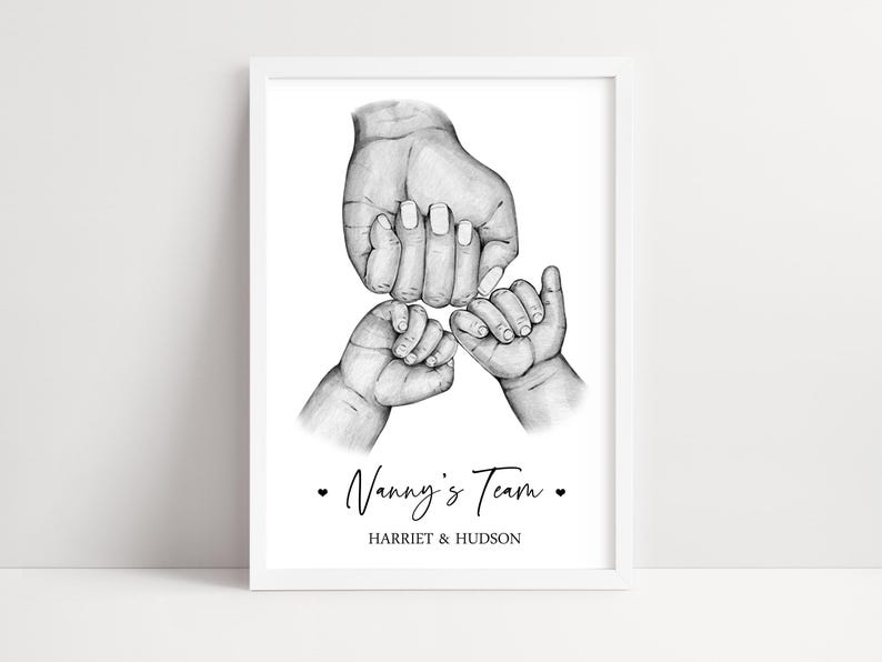 May include: A framed black and white pencil sketch print featuring one large hand holding two smaller hands. Below the illustration, the text reads Nanny's Team with a heart on each side, followed by the names HARRIET & HUDSON in capital letters.
