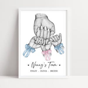 May include: A framed print featuring a black and white pencil sketch of one large hand holding three smaller hands. The smaller hands feature light blue and pink ribbon bows at the wrists. The text reads Nanny's Team, FINLEY, OLIVIA, BRODIE.