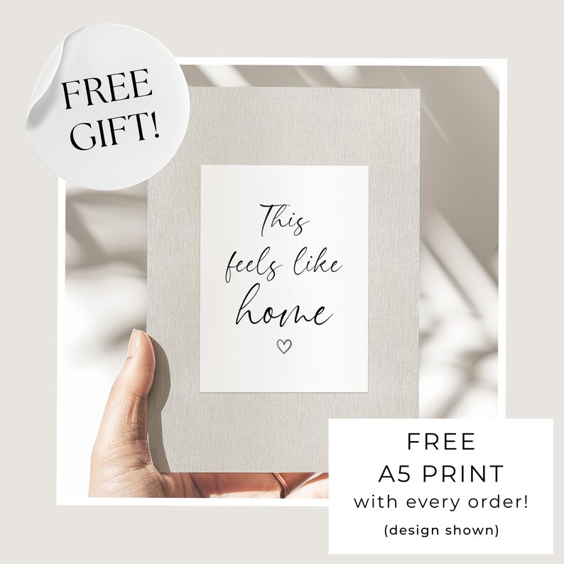 May include: A rectangular card with a light gray textured border and a white center panel featuring the black text This feels like home and a small heart icon. The card is held up against a neutral background with soft shadows cast across the surface.
