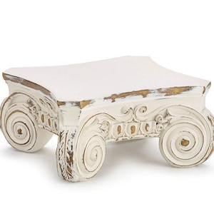 May include: A rectangular display stand featuring a white finish with distressed gold accents. The base is supported by four legs designed with prominent scroll-like volutes, reminiscent of an Ionic column capital, with intricate carved detailing.