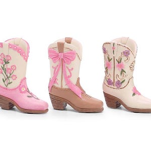 May include: Three decorative cowboy boots in a row. The boots are cream, pink, and brown with floral and bow accents. The boots have a small heel and are designed with a Western theme.