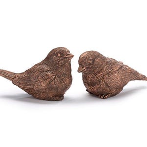May include: Two small, copper-colored bird figurines. The birds are facing each other, with one bird looking to the left and the other looking to the right. The birds are made of a textured material that resembles feathers.