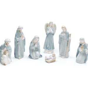 May include: A seven-piece porcelain nativity set featuring figures with a glossy finish in shades of soft blue, white, and pale peach. The set includes an angel, a kneeling figure, a standing figure, a baby in a manger, and three other figures.