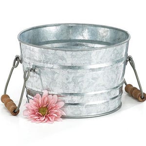 May include: A galvanized metal bucket with two wire handles and wooden grips. A pink flower rests near the base of the bucket. The bucket has a round shape and is made of corrugated metal.