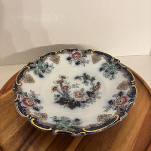 May include: A white porcelain plate with a floral design. The plate has a scalloped edge with a dark blue border and gold trim. The floral pattern includes red, pink, and green flowers.