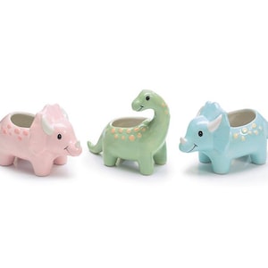 May include: Three ceramic dinosaur planters in pastel colors. A pink triceratops, a green brontosaurus, and a blue triceratops. Each planter has a round opening for plants and is decorated with small dots.