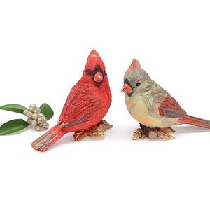 May include: Two decorative cardinal bird figurines. One is bright red with a black mask, the other is tan and gray. Both are perched on gold-colored leaf bases. A sprig of green leaves and berries is to the left.