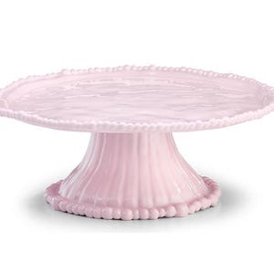 May include: A light pink cake stand with a round top and a pedestal base. The edge of the top and the base are decorated with small, round beads. The pedestal has vertical ribbed detailing. The stand is made of ceramic or glass.