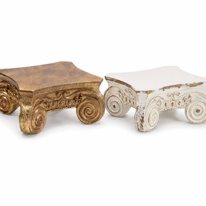 May include: Two rectangular display pedestals featuring ionic column capital designs with scrolled volutes. One pedestal has a metallic gold finish, while the other has a distressed white finish with gold accents visible on the carved details.