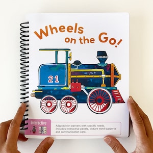 Puede incluir: Un libro colorido titulado "Wheels on the Go!" con una ilustración de un tren azul y rojo con el número 21 en el lateral. El libro es interactivo y adaptado para alumnos con necesidades específicas.