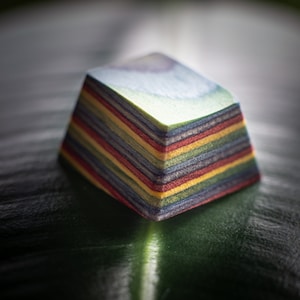 May include: A colourful, layered, geometric object with a rainbow pattern. The object is made of thin, stacked layers of wood in various shades of red, orange, yellow, green, blue, and purple. The object is sitting on a green leaf.