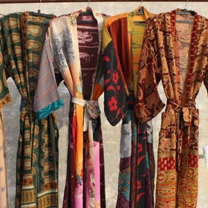 May include: A collection of colorful, patterned robes hanging on a brown metal rack. The robes feature a variety of designs, including paisley, floral, and abstract patterns, with a mix of colors like blue, green, red, and orange. Each robe has a matching belt.