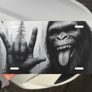 May include: Black and white license plate featuring a gorilla with its tongue out and a hand making the rock and roll gesture. The background shows a forest scene. The plate has four mounting holes.