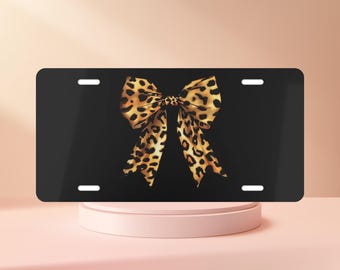 May include: A rectangular black license plate featuring a centered graphic of a large bow with a brown and black leopard print pattern. The plate includes four mounting slots, two positioned along the top edge and two along the bottom edge.
