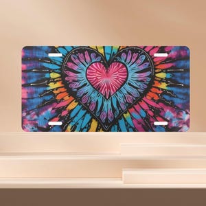 May include: A license plate featuring a tie-dye heart design. The heart is a vibrant mix of pink, blue, and black, set against a backdrop of swirling colors. The plate has four mounting holes.