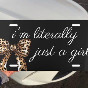 May include: Black license plate with the text "I'm literally just a girl" in white script. A leopard print bow is on the left side. The plate has four mounting holes.