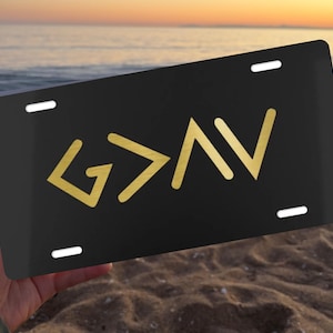 May include: A rectangular black license plate featuring the gold-colored text G > I V. The plate has four mounting slots, one in each corner, and a smooth, flat surface. The design is centered on the plate, displaying the letters and symbol clearly.