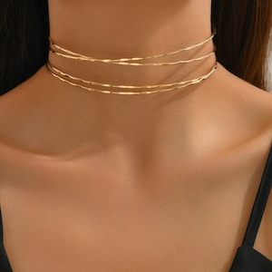 May include: A gold layered choker necklace with a twisted design. The necklace is worn by a person with a black top.