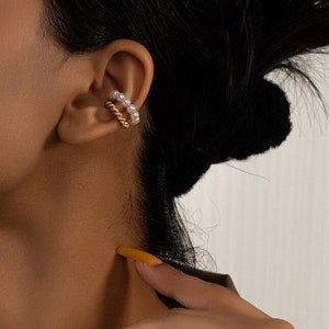 Puede incluir: Un par de pendientes de oro y perlas. Los pendientes están hechos de un alambre de oro trenzado con pequeñas perlas blancas unidas. Los pendientes se llevan en la parte superior de la oreja.