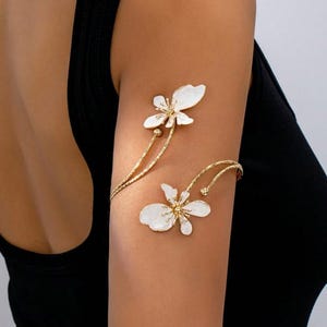 May include: Gold-tone arm cuff with two white flower designs. The floral details have gold centres and delicate petals. The cuff wraps around the upper arm, creating an elegant and unique accessory.