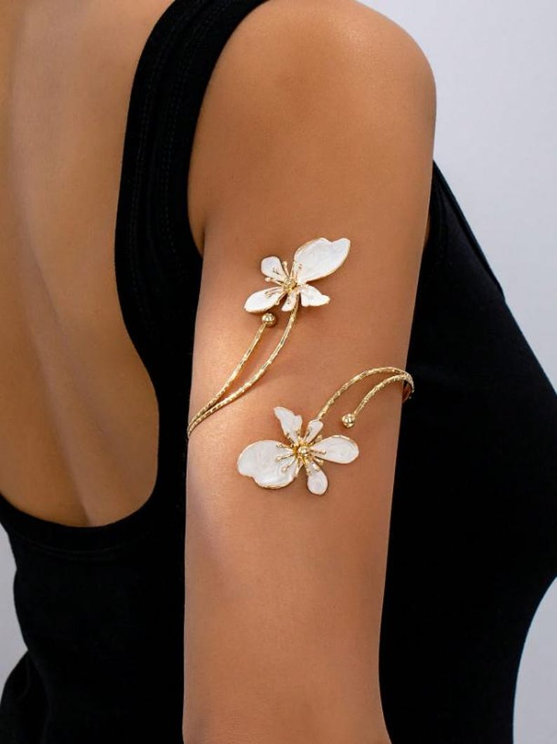 May include: Gold-tone arm cuff with two white flower designs. The floral details have gold centres and delicate petals. The cuff wraps around the upper arm, creating an elegant and unique accessory.