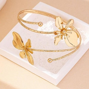 May include: Gold-coloured arm cuff with two stylized flower designs. The cuff wraps around the arm with a delicate, open design. The flowers have detailed petal shapes, adding a touch of elegance to the jewellery.
