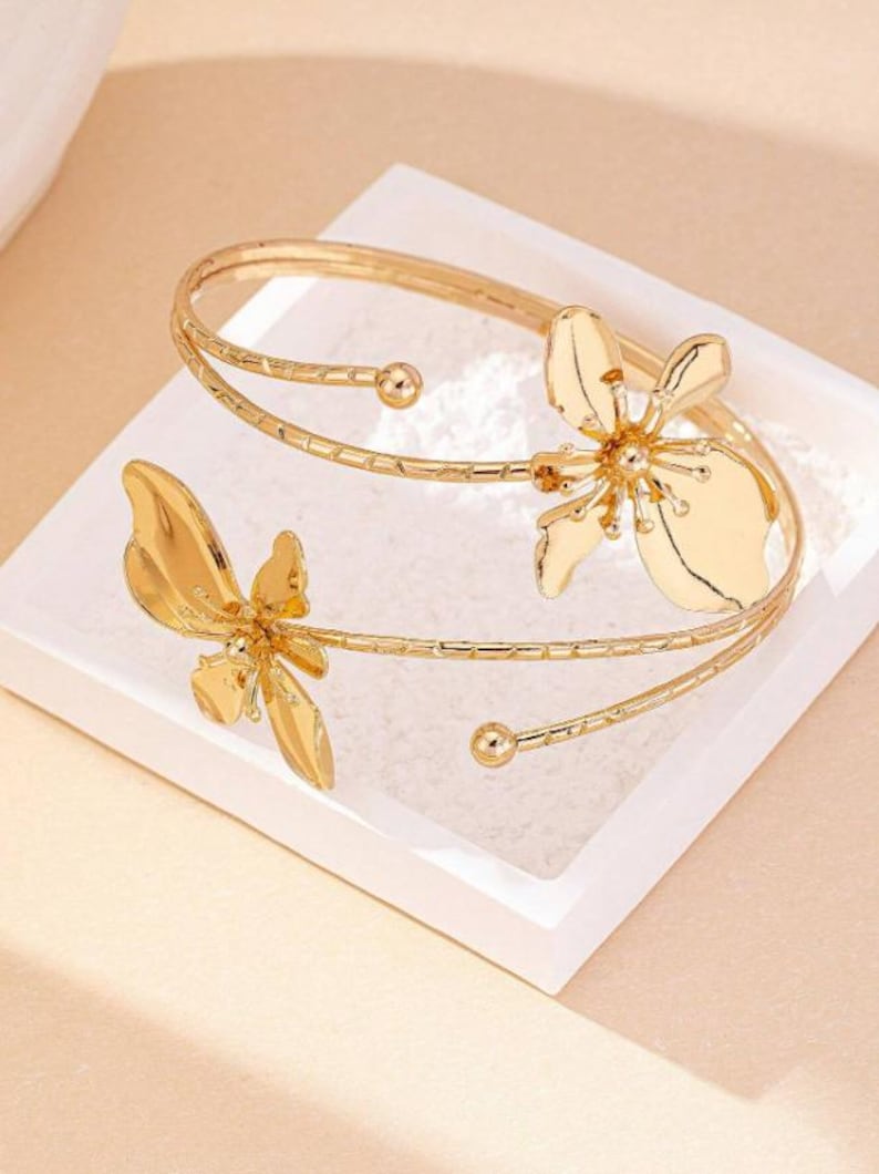 May include: Gold-coloured arm cuff with two stylized flower designs. The cuff wraps around the arm with a delicate, open design. The flowers have detailed petal shapes, adding a touch of elegance to the jewellery.