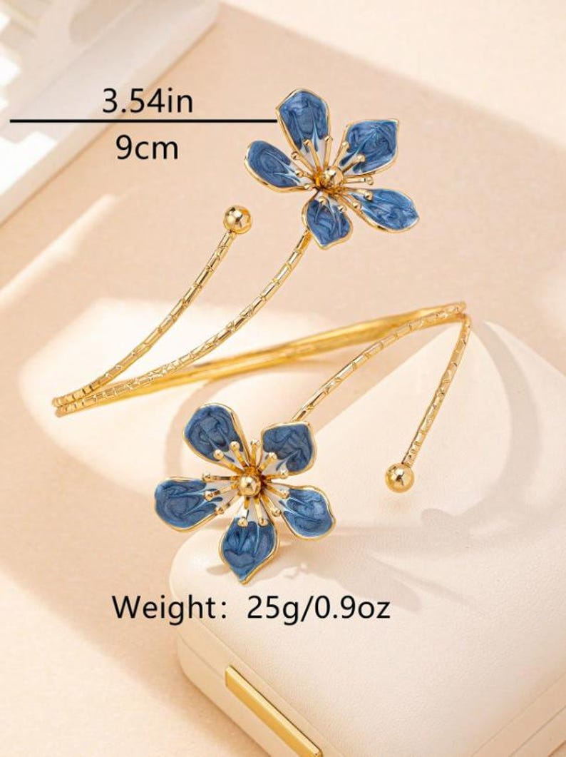 May include: Gold-coloured arm cuff with two blue flower designs. Each flower has five petals with a blue enamel finish. The cuff is 9 cm long and weighs 25g.