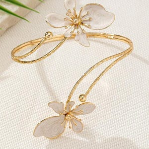 May include: A gold-coloured arm cuff with two white flower designs. The flowers have detailed petal designs with gold centres. The cuff has a delicate, open design, with a curved shape that wraps around the arm.