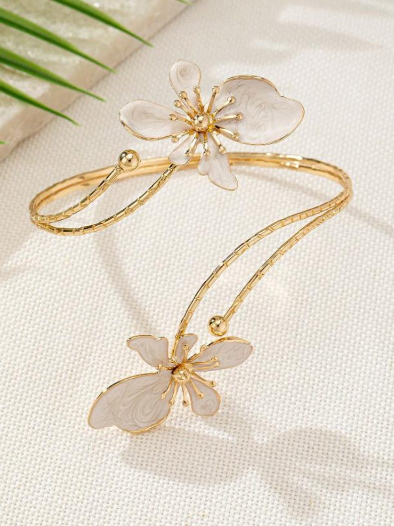 May include: A gold-coloured arm cuff with two white flower designs. The flowers have detailed petal designs with gold centres. The cuff has a delicate, open design, with a curved shape that wraps around the arm.