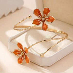 May include: Gold-coloured arm cuff with two orange flower features. The flowers have a detailed design with a central gold-coloured bead. The cuff has a delicate, open design, with a small gold bead at each end.