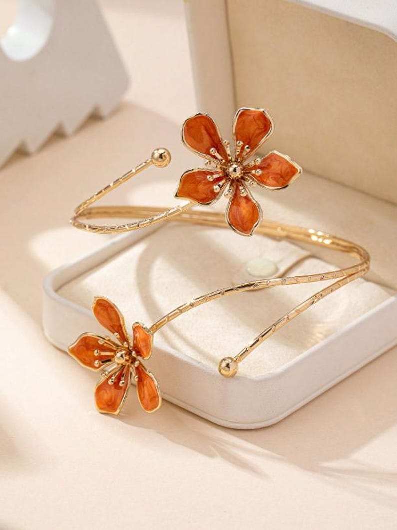 May include: Gold-coloured arm cuff with two orange flower features. The flowers have a detailed design with a central gold-coloured bead. The cuff has a delicate, open design, with a small gold bead at each end.