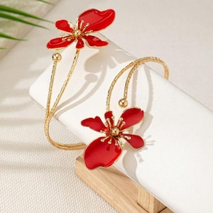 May include: Gold-coloured arm cuff with two red flower designs. The flowers have a glossy finish and detailed centres. The cuff is displayed on a white surface, showcasing the jewellery's elegant design. The floral design adds a touch of nature-inspired beauty.