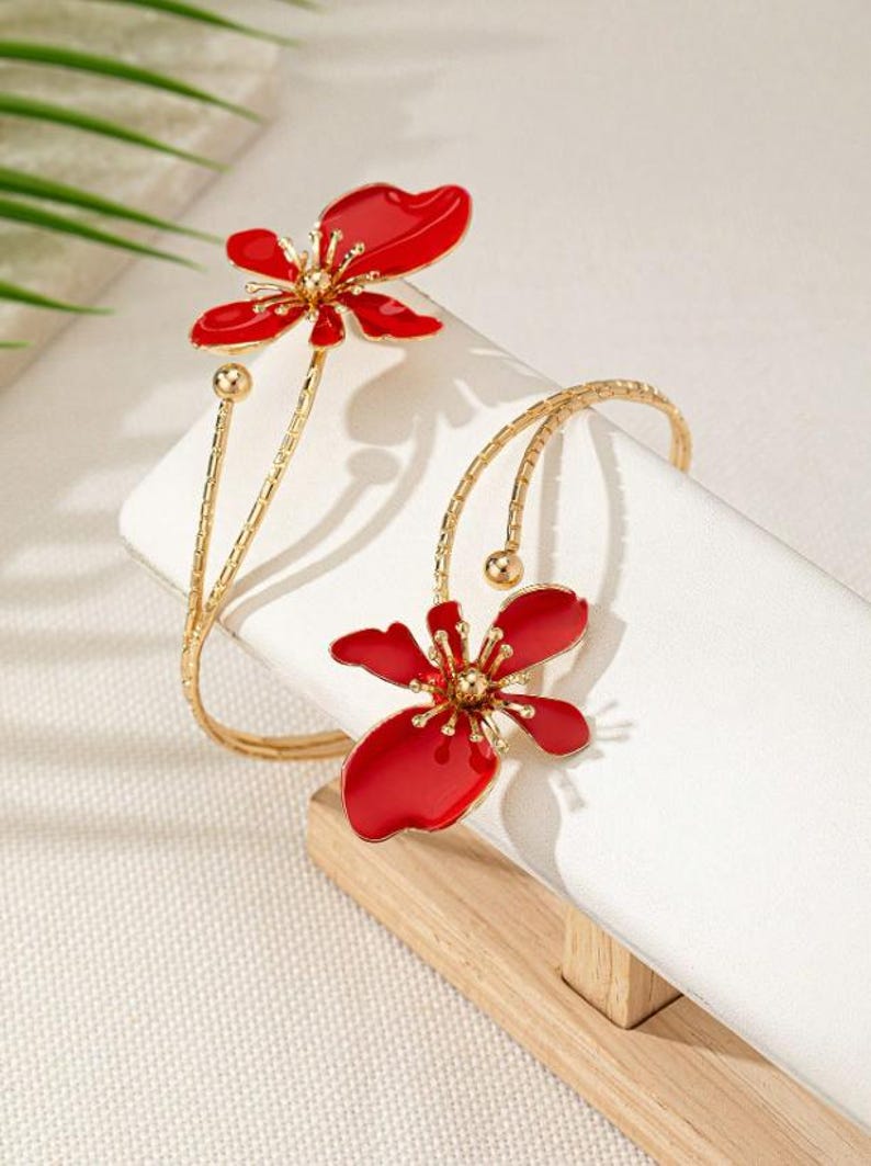 May include: Gold-coloured arm cuff with two red flower designs. The flowers have a glossy finish and detailed centres. The cuff is displayed on a white surface, showcasing the jewellery's elegant design. The floral design adds a touch of nature-inspired beauty.