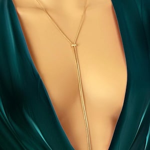 May include: A gold-coloured necklace with a Y-shaped design. The necklace has a delicate chain that splits into two strands, creating a long, elegant drop. The necklace is shown against a dark green background.