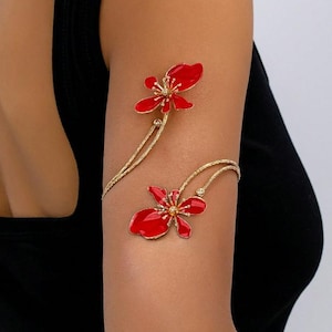 May include: Gold-coloured arm cuff with two red flower designs. The flowers have detailed centres and are connected by delicate, curved gold lines. The jewellery is worn on the arm, showcasing the floral design.