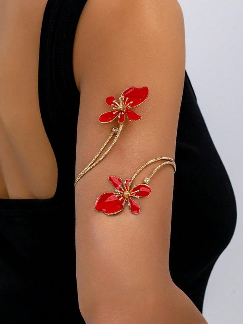 May include: Gold-coloured arm cuff with two red flower designs. The flowers have detailed centres and are connected by delicate, curved gold lines. The jewellery is worn on the arm, showcasing the floral design.