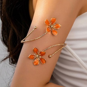 May include: Gold-tone arm cuff with two orange flower designs. The floral accents have a detailed centre and are connected by delicate, curved gold-tone bands. The arm cuffs are designed to be worn on the upper arm.